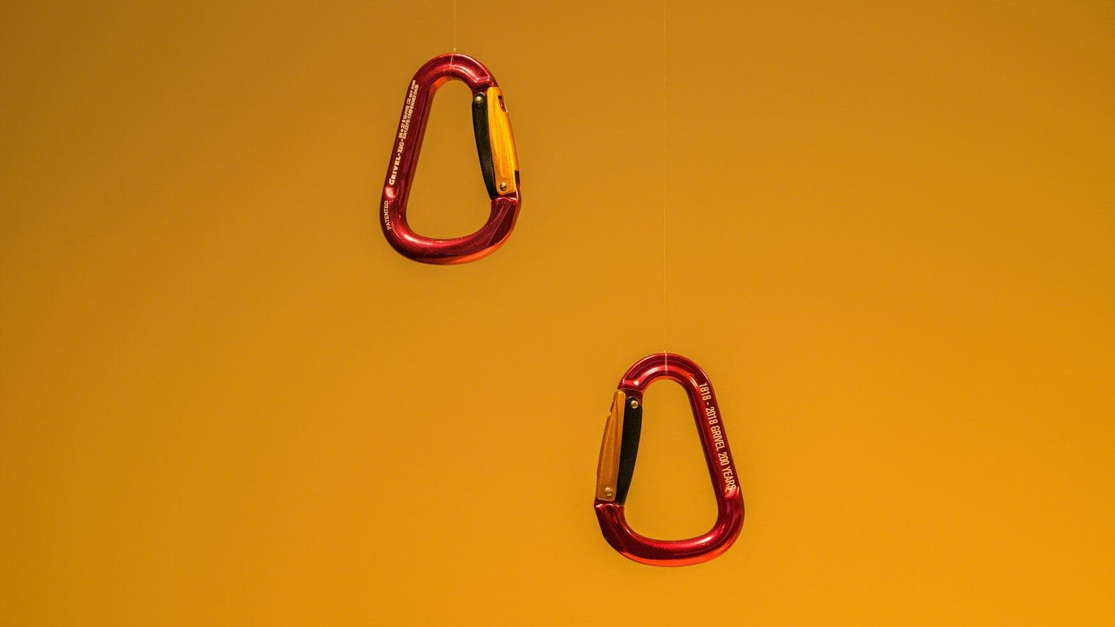 Dois mosquetões de alumínio anodizado em vermelho pendurados contra um fundo amarelo