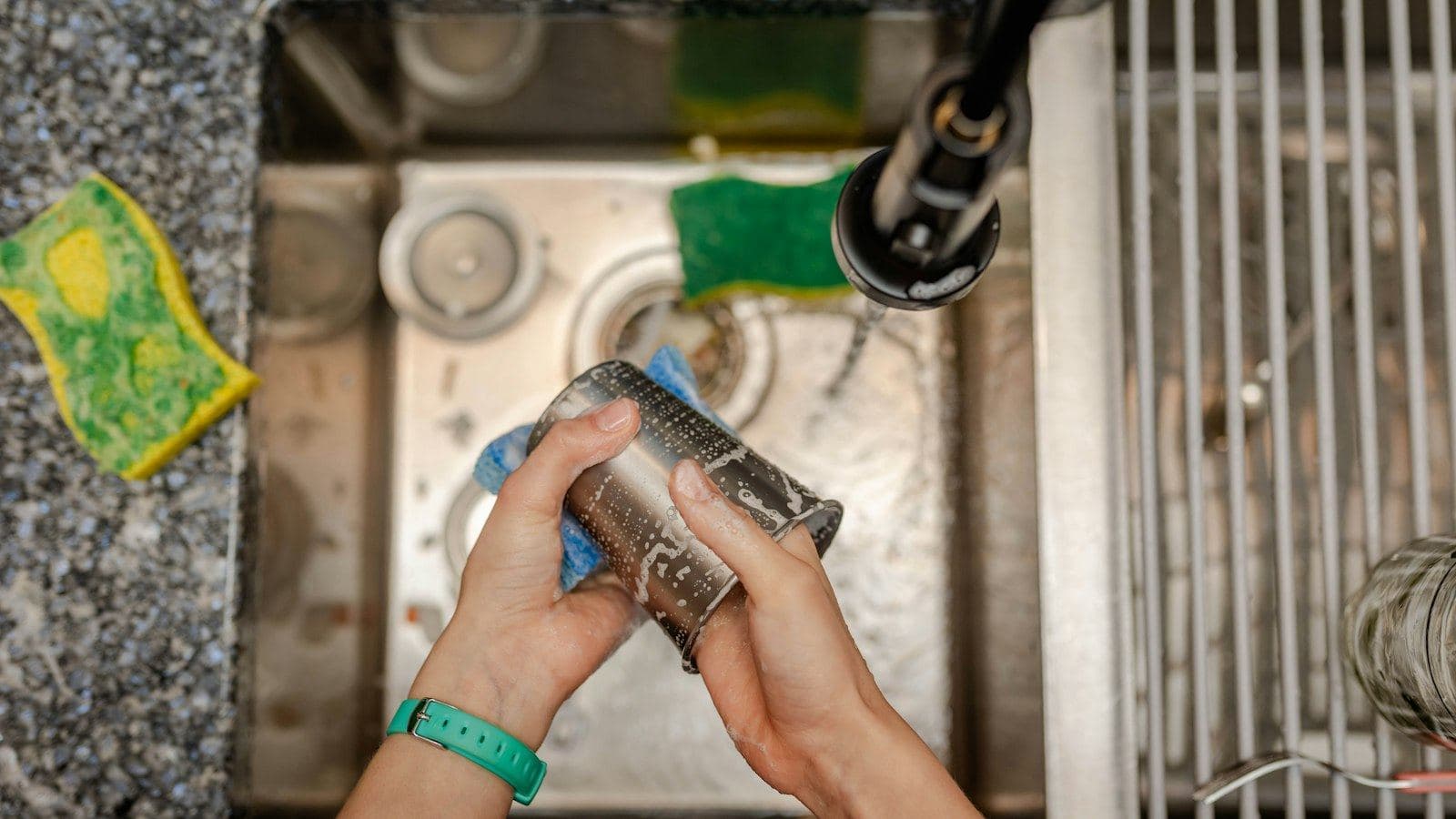 Mãos lavando com esponja um copo de aço inox dentro de uma pia também de inox